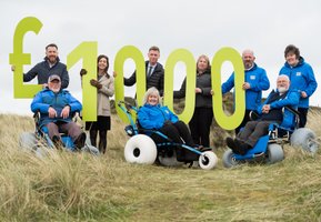 Beach wheelchairs and volunteers holding £1000 sign in the sand dunes
